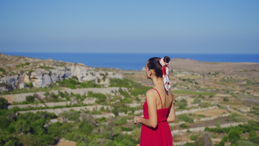 A young slender brunette woman stands on a cliff at a height and considers nature, shows her hand. Back view