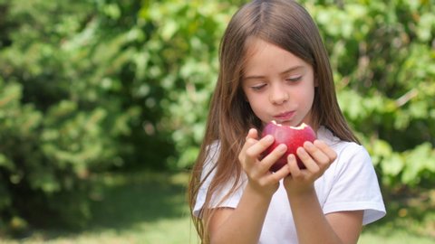 Cute little girl bites and eats a fresh red apple while standing in the garden. Chews and smiles. Healthy tasty food concept.