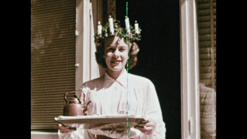 1950s: Group of people in a line ski down slope. People ski down slope. People ski passed trees. Woman walks through doorway wearing wreath of candles on head and carrying a platter.