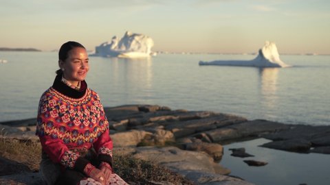 Tilt up: Smiling woman in national costume looking at Disko Bay while sitting on rock