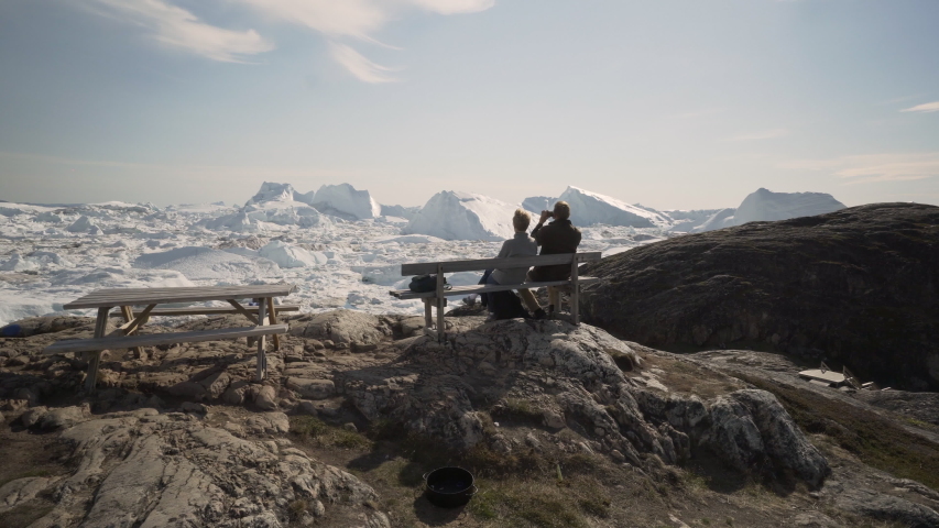 Slow Motion: Two Tourists Sit on a Wooden Bench on Rocky Outcrop While Taking Photos of Ice Fjord - Disko Bay, Greenland