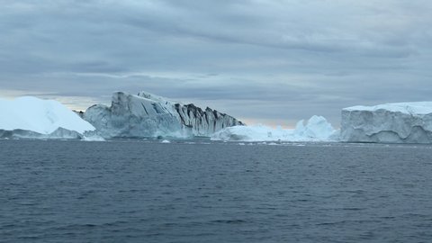 Pan: Massive Icebergs Jutting Out From the Water in Disko Bay Before Cloudy Sky
