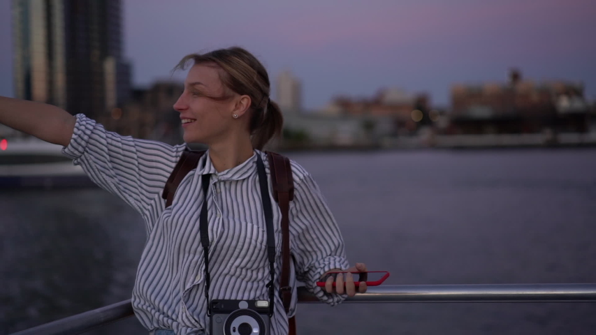 Slow motion effect of beautiful hipster girl enjoying voyage trip on east river for making sightseeing on water, young Caucasian female tourist with instant camera and mobile phone exploring city