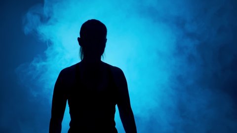 Silhouette of boxer woman showing biceps before sparring on dark background of blue smoke.