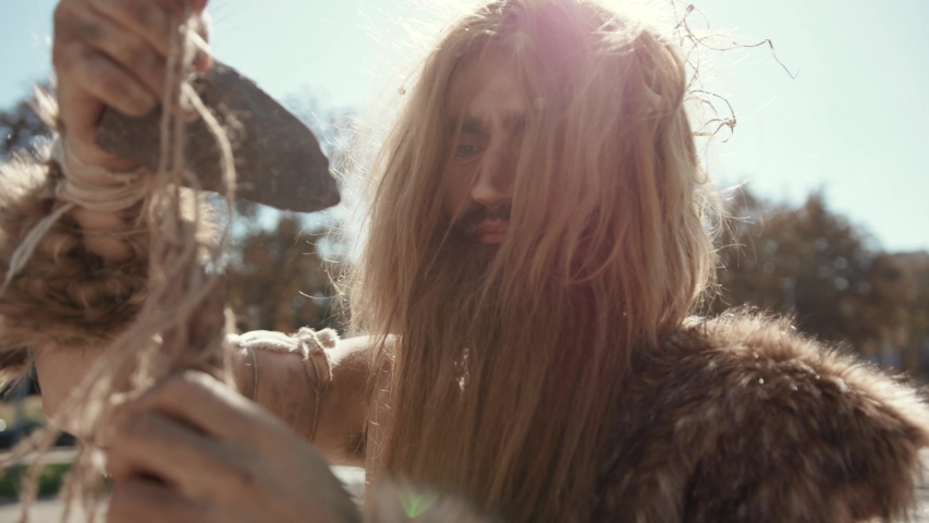 Close-up of neanderthal savage making a stone-tipped spear creating hunting tool staying in modern city street. Wild homo sapiens. Evolution.