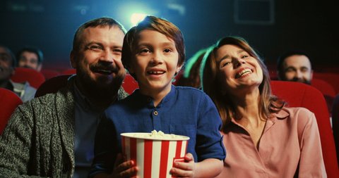 Portrait of teh happy small cute boy with popcorn in hands sitting in the cinema between cheerful parents and they laughing as watching comedy movie.
