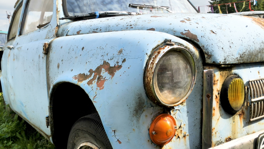 The white car with the rust on the body of it parked along side other abandoned old cars