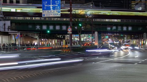 Timelapse zoom out Kobe public transport traffic on above-ground city rail overpass and bus stop under metro station with people crowd and cars flow along metropolis highway at night rush hour
