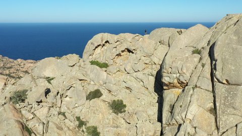 (Drone Footage) View from above, stunning aerial view of a person on the top of a granite mountain. Cala Coticcio also known as Tahiti in the background. La Maddalena Archipelago, Sardinia, Italy. 
