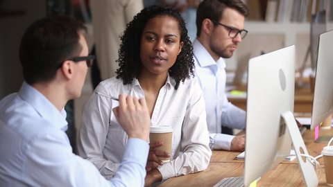 Diverse workers working in coworking, focus on mixed race woman employee receive instructions help with task from caucasian mentor, colleagues discuss project sit at workplace near pc in modern office