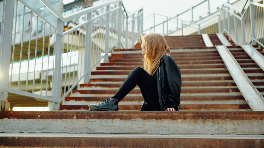 Young fashion model in rock star look posing on staircase. Teenager girl in black leather jacket sitting on stairs and looking to camera