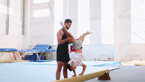 A young boy athlete is making a handstand on the gymnastic beam and pushing up headfirst in the sport center. A beard fit partner man is lifting a teenager by his legs. Side view.