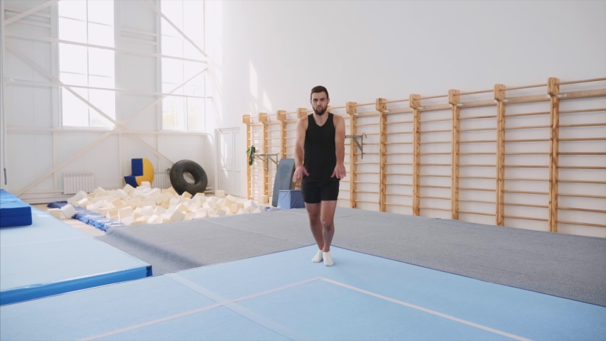 A male athlete is making four back flips at once in front of the camera and lands on feet in the gym in black shorts and T-shirt, sport equipment at the background, steadicam, front view, slow motion. Royalty-Free Stock Footage #1038205721