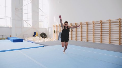 A male athlete is making four back flips at once in front of the camera and lands on feet in the gym in black shorts and T-shirt, sport equipment at the background, steadicam, front view, slow motion.