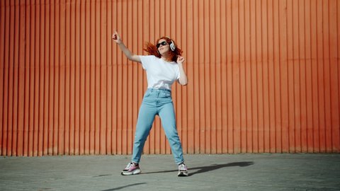 Slow motion of happy teenager in headphones and sunglasses dancing singing outdoors in city street having fun alone. Lifestyle, leisure and people concept.