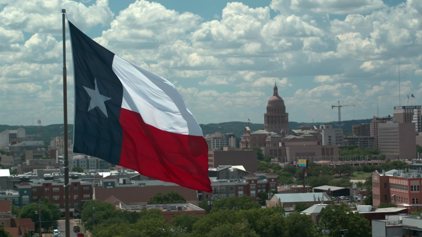 A large Texas flag is waving, captured with a static aerial camera. The background is downtown Austin and the Texas Capital building. The camera used a long lens to give the image the classic "helicop Royalty-Free Stock Footage #1038219056