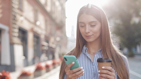 Attractive Fashionable Young Girl with Headband Using her Smartphone and Smiling Holding a Paper Coffee Cup Standing at Old City Background Close Up