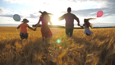 Young couple of parents with two children holding hands of each other and running through wheat field at sunset. Happy family jogging among barley meadow and enjoying nature together. Slow motion