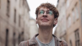 Close Up of Young Smiling Hipster Guy in Glasses Wearing Checked Jacket in Earphones with Bag Walking, Looking around at Old City Street. Student, Tourist Concept. Traveling. - Powered by Shutterstock - Get 15% off with code: PIKWIZARD15