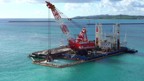 Huge excavator on a floating platform in the blue water of Pacific Ocean extracts sand from the bottom and loads onto a barge against the background of clouds and tropical islands
deepening of seabed.