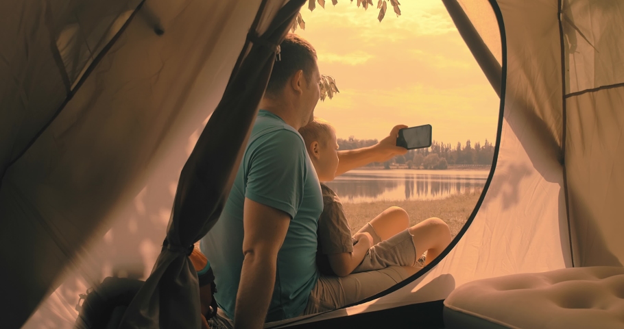 Happy traveler father and son sitting near tent camp around mountains lake and shoot with her phone camera under sun light enjoying the leisure and freedom. 4K slow motion video

