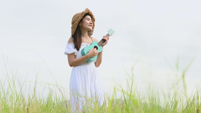 Asian happy woman in white dress playing ukulele guitar in green meadow field with mountaing background on summer. Musician beauty woman relax with musical instrument. Lifetyle vintage in countryside