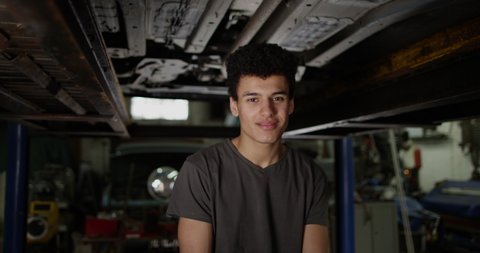 4K Apprentice garage mechanic standing under vehicle ramp & smiling at camera
