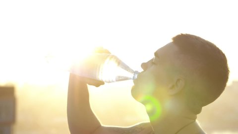 Energy sports drink - athlete drinking from water bottle on training. Attractive man taking break and drinks water from bottle outdoors at sunset. Beauty sunset time
