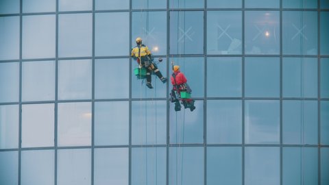 Two men workers in red and yellow work clothes cleaning the exterior windows of a business skyscraper - industrial alpinism