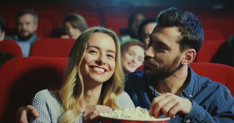 Portrait of the happy romantic Caucasian couple, beautiful blond woman and handsome man hugging and smiling while eating a popcorn and watching a film.