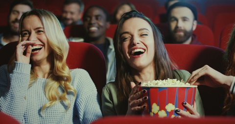 Close up of the three Caucasian pretty and joyful female best friends sitting and laughing loud in the cinema while holding popcorn and watching a comedy film.