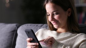 Happy woman uses mobile phone sitting on a couch in the night at home