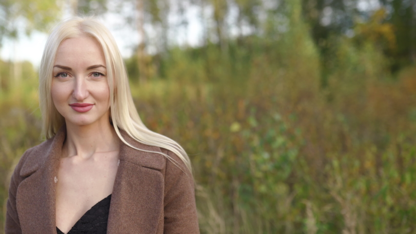 Blonde woman portrait in outside autumn park with golden and green forest in the background - Wearing a brown coat