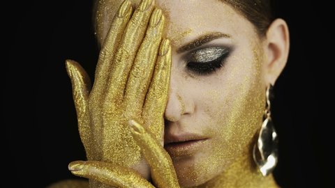 Girl face with golden make-up  in the studio on  black background.  Gold paint on the face and fingers. Hand covers face Gold earrings, gold eye shadow, clear skin. 