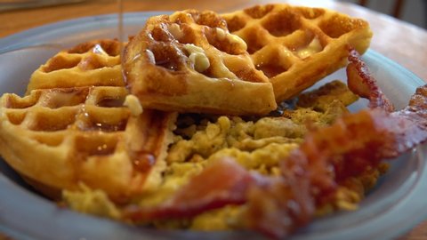 Pouring syrup over waffles on plate with bacon and scrambled eggs for breakfast