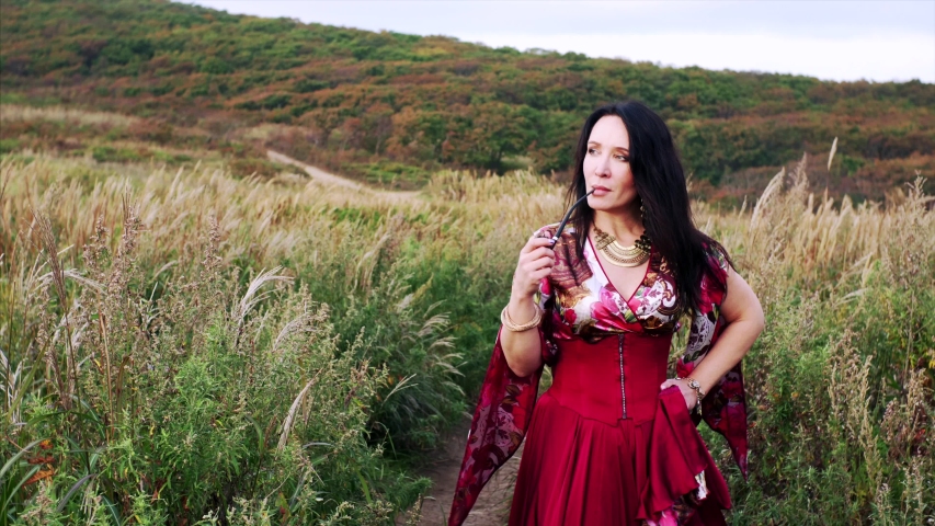 Portrait of attractive gipsy woman in colorful dress smoking pipe with light smile and walking along the path in the field with high grass. Summer
