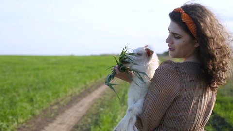Young woman with curly brown hair holding a little white goat on the background of green field and country road. Beautiful plus-size model posing. Friendship of people and animals. Vegetarian concept.