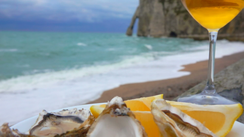 Close-up of a plate full of fresh oysters and a glass of white wine against the ocean on a cloudy day in Etretat, France