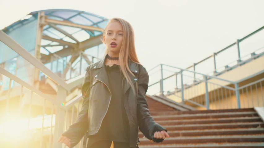 Stylish girl in leather jacket dancing on stairs outdoor. Low angle view of fashionable teenage girl with bright makeup wearting black leather jacket and dancing on steps in urban cityscape sunset.