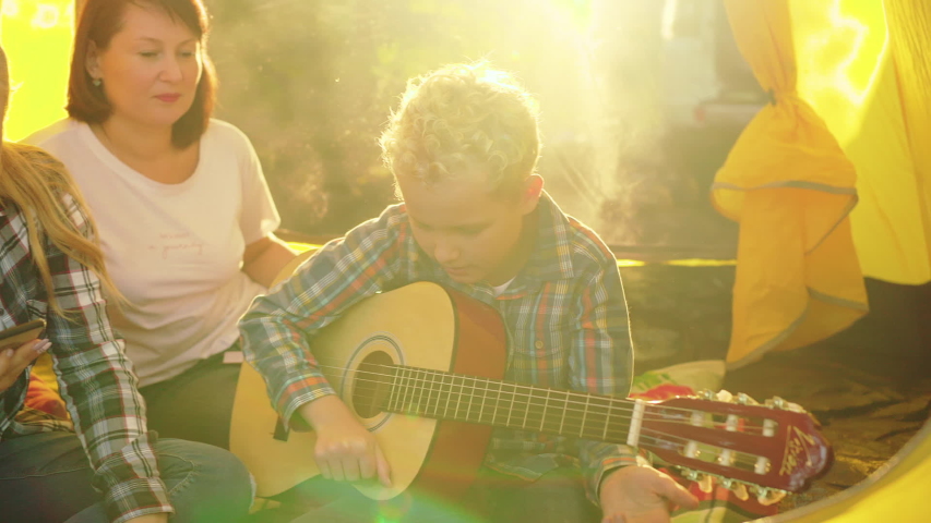 Mother with son and daughter enjoying guitar in tent. Middle aged woman talking, boy playing guitar and girl using smartphone while sitting together in tent. Camping concept