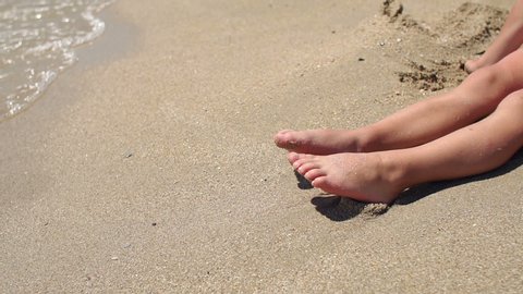 Close-up of a little boy's feet on the beach on the sand. The child sits on the warm sand on the beach, close-up. Slow motion.