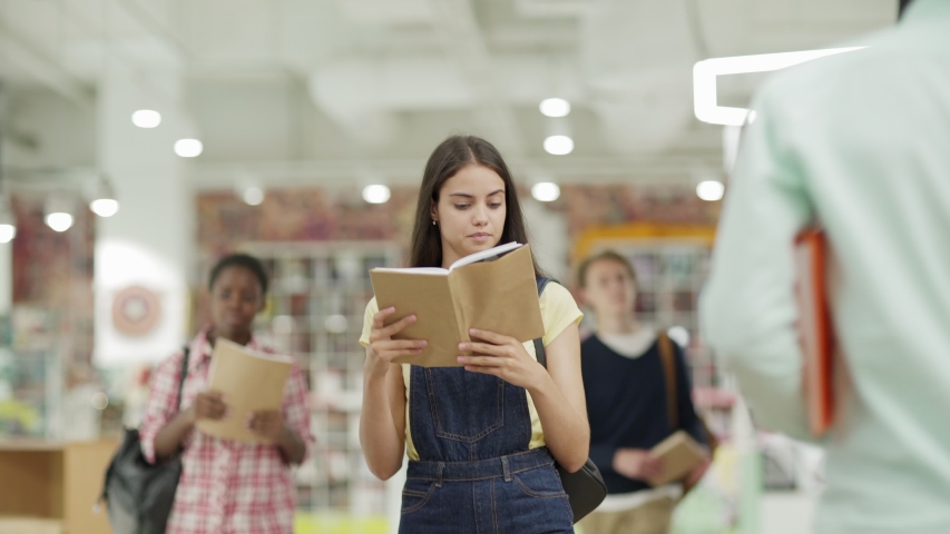 Waist up shot of intelligent college girl walking along shelves in library and reading book, male friend joining her. Two students talking 