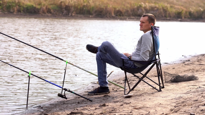 The guy is fishing while sitting on a chair by the lake. Warm summer day, sunny weather.