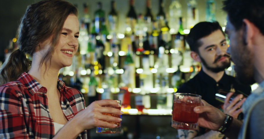 Close up of the young pretty Caucasian girl smiling while spending time with her boyfriend and talking with bartender while he serving and doing cocktail ar the bar.