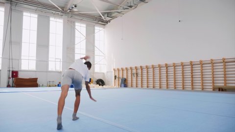 A teenager boy in a white T-shirt and grey shorts is doing a cartwheel and a triple back flip and lands on feet in gym on the blue sport mats, steadicam shot.