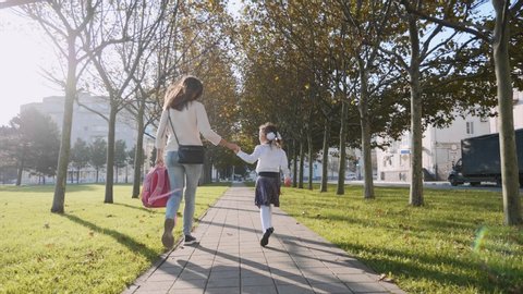 Young woman and little girl in school uniform are holding hands and running skipping along the trees in the park at sunny autumn weather. Family late for school concept, steadicam shot back view.