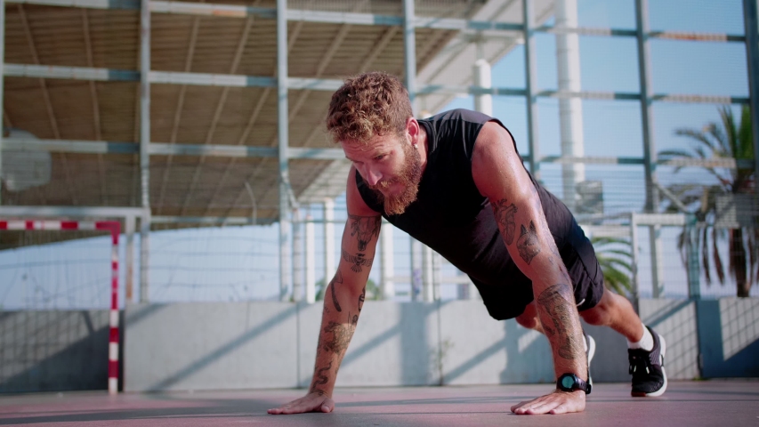 Strong energetic Caucasian bearded man with tattooed hands pushing-up at summer morning, leaning on play ground self-confidence man healthy sport lifestyle dressed in black shirt shorts and sneakers