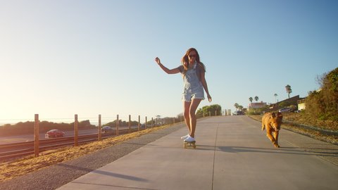 Young happy smiling girl skateboarding with golden retriever puppy at sunset