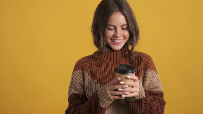 Pretty smiling brunette girl in cozy sweater dreamily smelling coffee on camera over yellow background