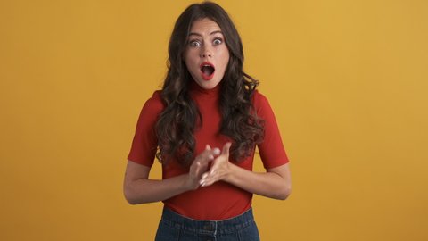 Attractive surprised brunette girl in red top amazedly looking in camera over yellow background. Wow expression